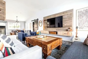 A modern living room with a fireplace, a large TV mounted on a wooden wall, a sofa, a coffee table, and a guitar. - Yucca Valley Oasis: Pool, Spa & Game Room Retreat (Yucca Valley)
