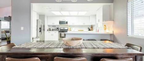 A modern dining area with a dark wood table and brown chairs, a chandelier overhead, and a view into a kitchen with white cabinets.