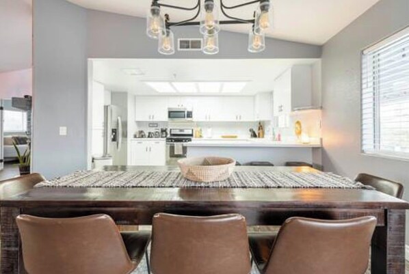 A modern dining area with a dark wood table and brown chairs, a chandelier overhead, and a view into a kitchen with white cabinets.