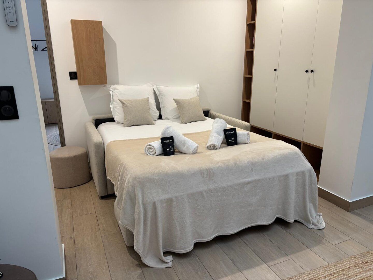 Une chambre d’hôtel moderne comprenant un lit, une table de chevet, une armoire et un petit pouf rond.