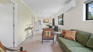 A modern living room with a green sofa, a brown leather chair, and a small table with a plant. There is a kitchen area in the background with a refrigerator and a window. - Birch Cottage - Greytown Holiday Home (Greytown)