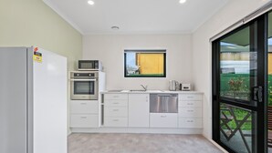A compact kitchen with a built-in oven, microwave, and dishwasher, a window with a view of a yellow building, and a sliding glass door leading to an outdoor area. - Birch Cottage - Greytown Holiday Home (Greytown)