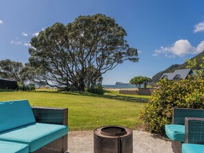 A patio area with wicker furniture, a large tree, and a view of the sea and hills. - Beach Haven Bonanza - Pauanui Holiday Home (Pauanui)