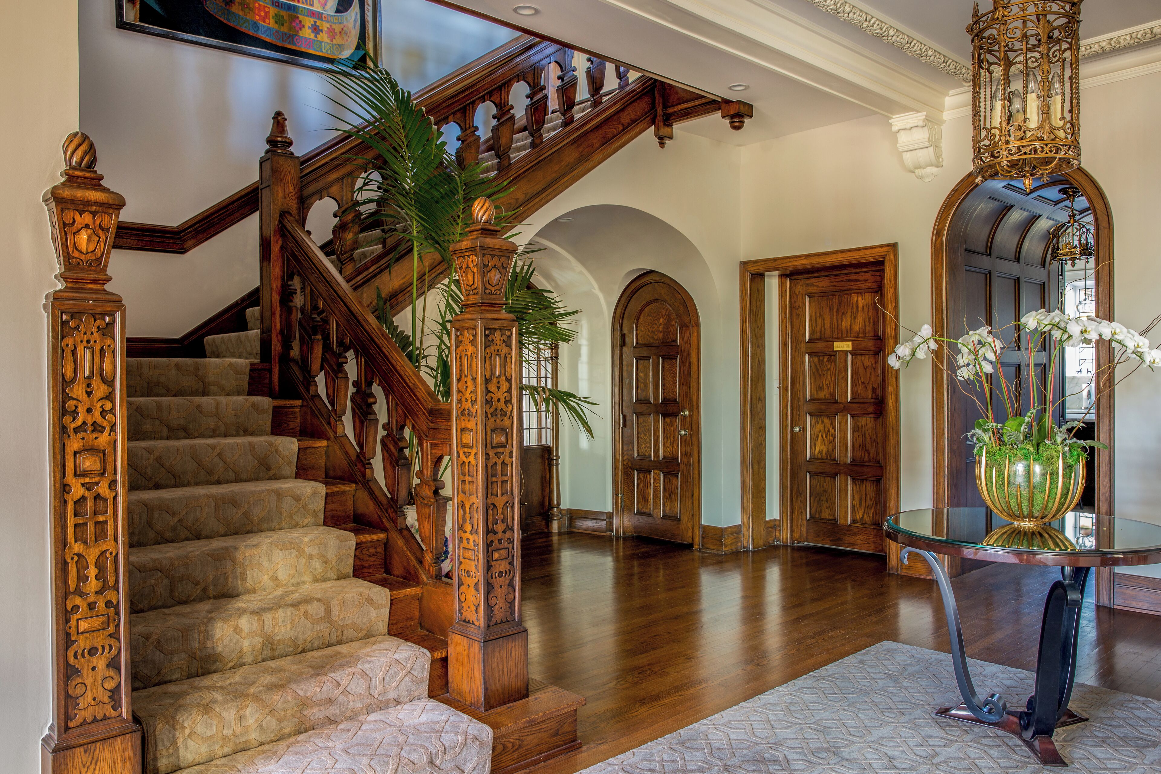 Una imponente escalera con detalles de madera ornamentados, una mesa de vidrio con una planta y un piso de madera pulida.