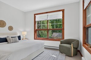 Une chambre avec une grande fenêtre, un lit, une chaise et une table de chevet avec une lampe.
