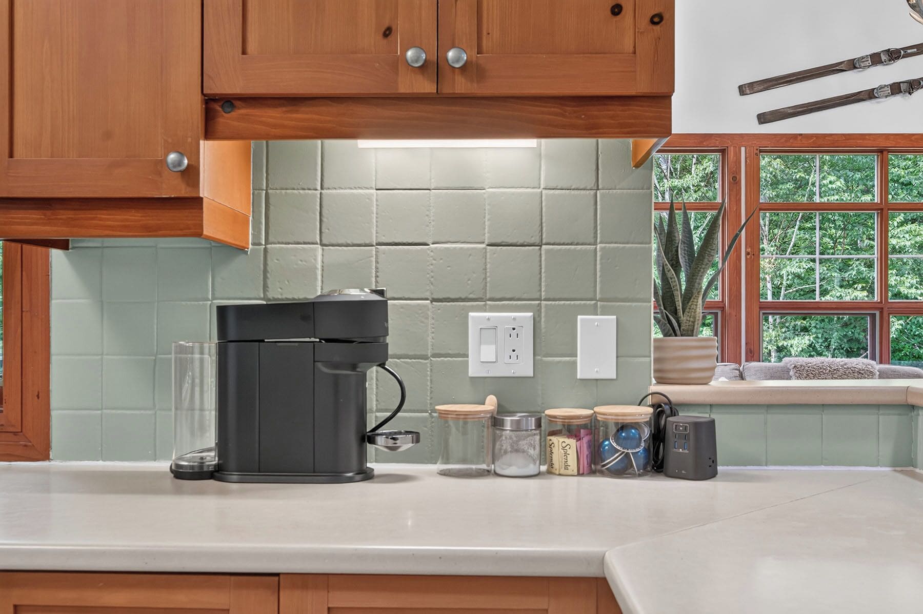 A kitchen with wooden cabinets, a coffee maker, jars, and a window with a view of greenery.