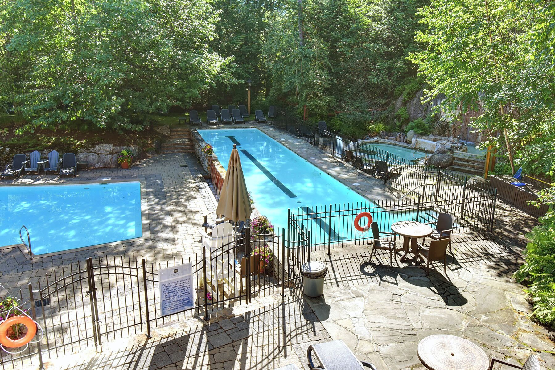 A rectangular outdoor swimming pool with a stone deck, surrounded by a metal fence. There are lounge chairs and umbrellas along the poolside.