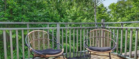Two wicker chairs with cushions on a wooden deck, overlooking a lush green forest.