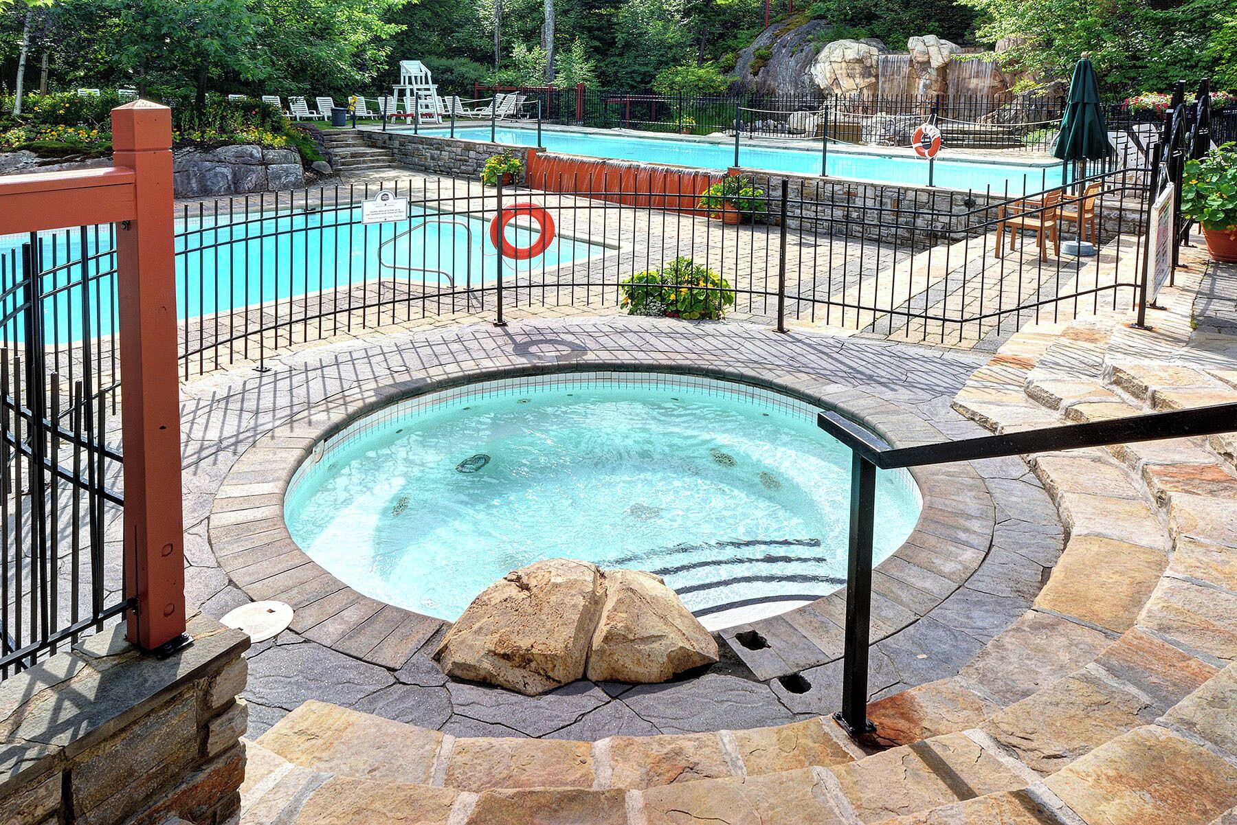 Un bain à remous entouré de pierres, une piscine avec une cascade et un espace clos aménagé avec des chaises longues.