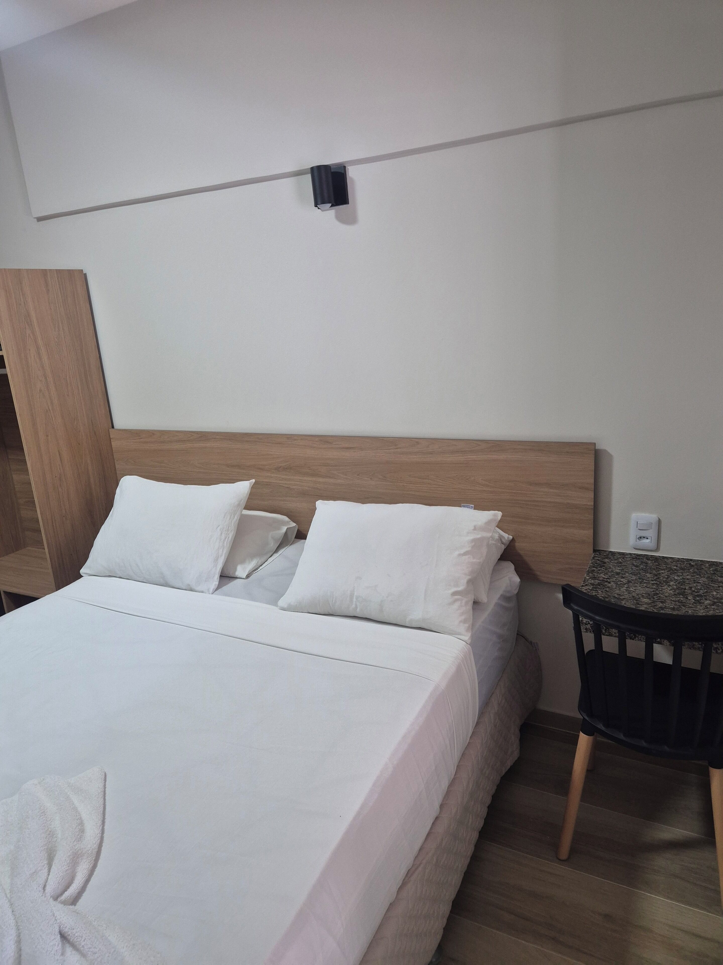 A neatly made bed with white linens, a wooden headboard, and a small table with a chair beside it.