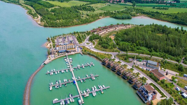 An aerial view of a marina with boats docked, surrounded by greenery and a small island. - Luxuriose Ferienwohnung am Stormthaler See (Grossposna)