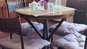 A round wooden table with a woven placemat, surrounded by cushioned chairs with a woven backrest. A small potted plant with purple flowers is on the table. - Sheiton Búzios (Búzios)