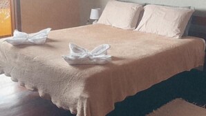 A neatly made bed with a beige bedspread and pillows, a bedside table with a lamp, and a window with blinds. - Sheiton Búzios (Búzios)