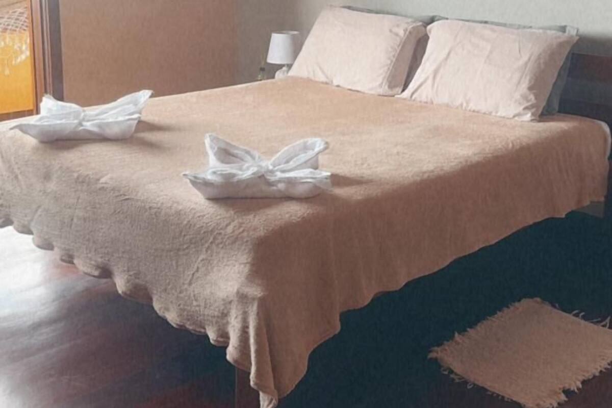 A neatly made bed with a beige bedspread and pillows, a bedside table with a lamp, and a window with blinds.
