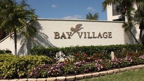 A sign for 'Bay Village' with a bird logo, surrounded by a hedge and flowering plants. - Charming 2b-2b condo in beautiful Fort Myers Beach- 10 min walk to the beach .  (Fort Myers Beach)