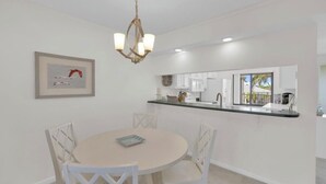 A dining area with a round table and white chairs, a kitchen with white cabinets, and a hanging light fixture. - Charming 2b-2b condo in beautiful Fort Myers Beach- 10 min walk to the beach .  (Fort Myers Beach)