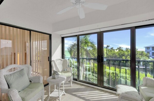 A balcony with a view of greenery and buildings, white wicker furniture, and a ceiling fan.