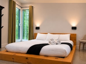 Une chambre moderne avec un lit en bois, du linge de lit blanc et une vue sur la verdure par la fenêtre.