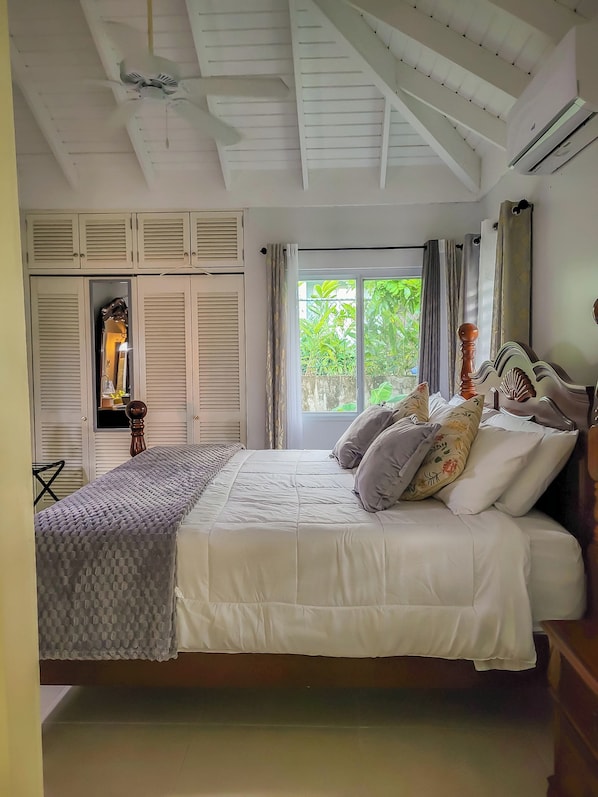 A bedroom with a large bed, a ceiling fan, an air conditioning unit, a window with curtains, and a wardrobe with louvered doors.
