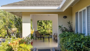 Een overdekt terras met buitenmeubilair, omgeven door weelderige begroeiing en een betegelde vloer.