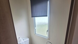 A compact bathroom with a toilet, a window with a blind, and a small sink. - Steve Caravan Sleeps 6 With Free Parking (Rhyl)
