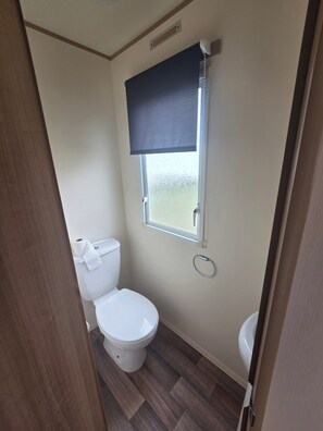 A compact bathroom with a toilet, a window with a blind, and a small sink. - Steve Caravan Sleeps 6 With Free Parking (Rhyl)