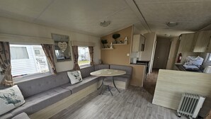 A compact RV interior with a sofa, a small dining table, a kitchenette, and a window with curtains. - Ruby a Modern Caravan in Rhyl (Rhyl)