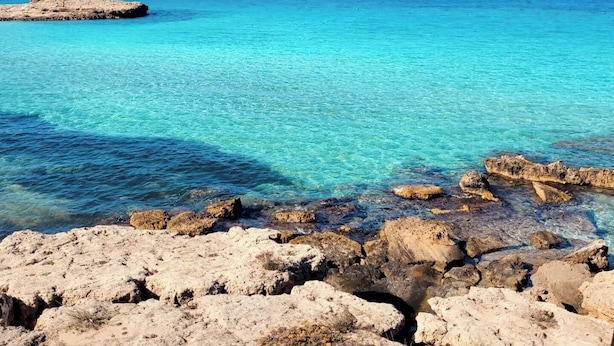 Eine felsige Küstenlinie mit klarem, türkisblauem Wasser und einer in der Ferne liegenden Felsenformation.