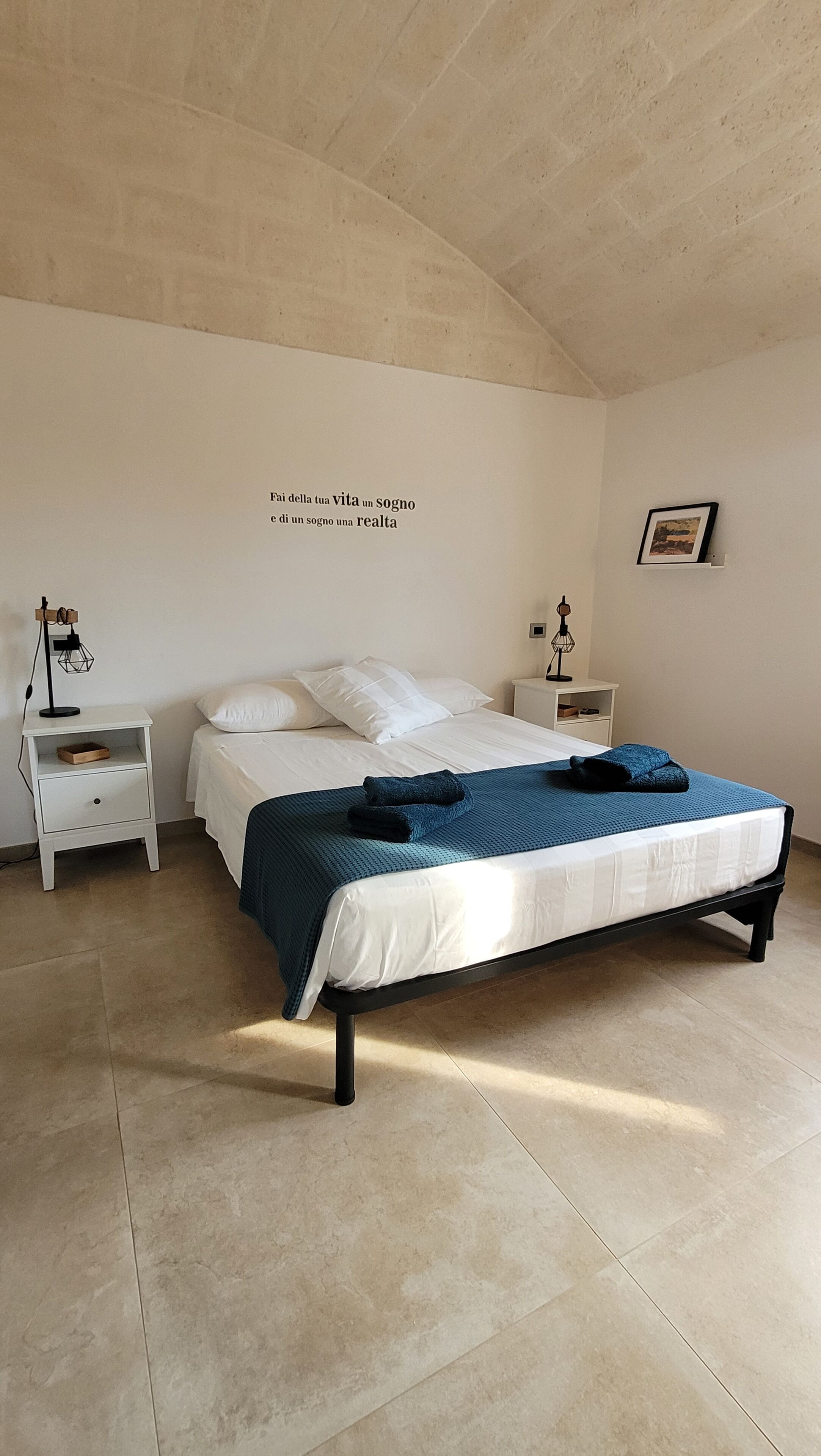 A bedroom with a bed, two bedside tables, a wall-mounted shelf, and a framed picture.
