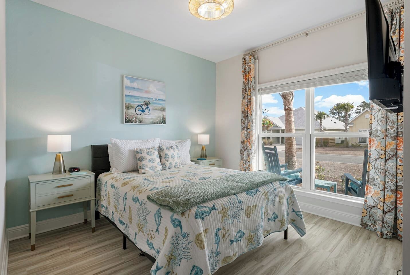 A bedroom with a bed, nightstands, a lamp, a painting, a window with curtains, and a view of the outside.