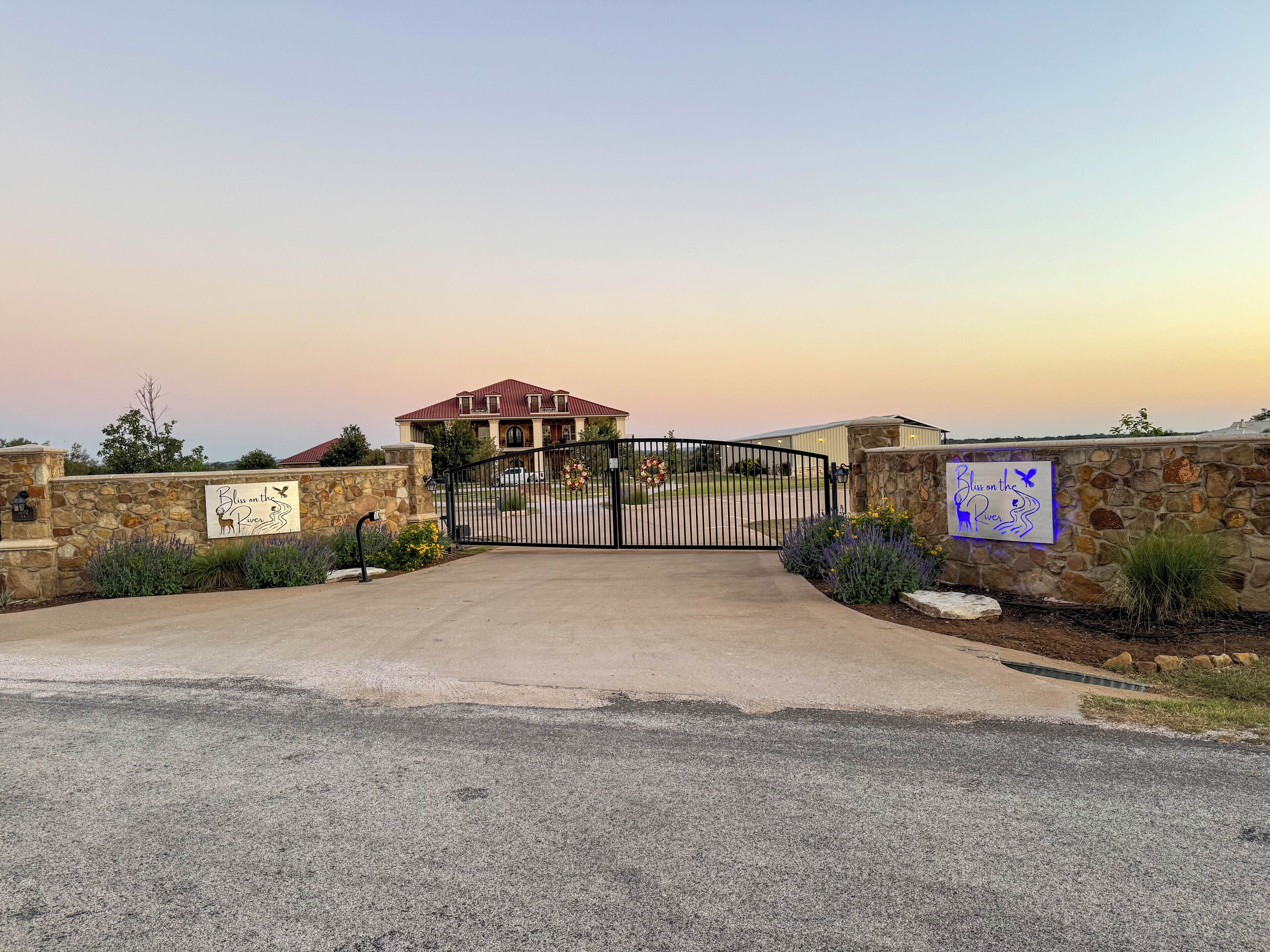 A gated entrance to a property with a stone wall, a metal gate, and a sign that reads "Bless on the River."