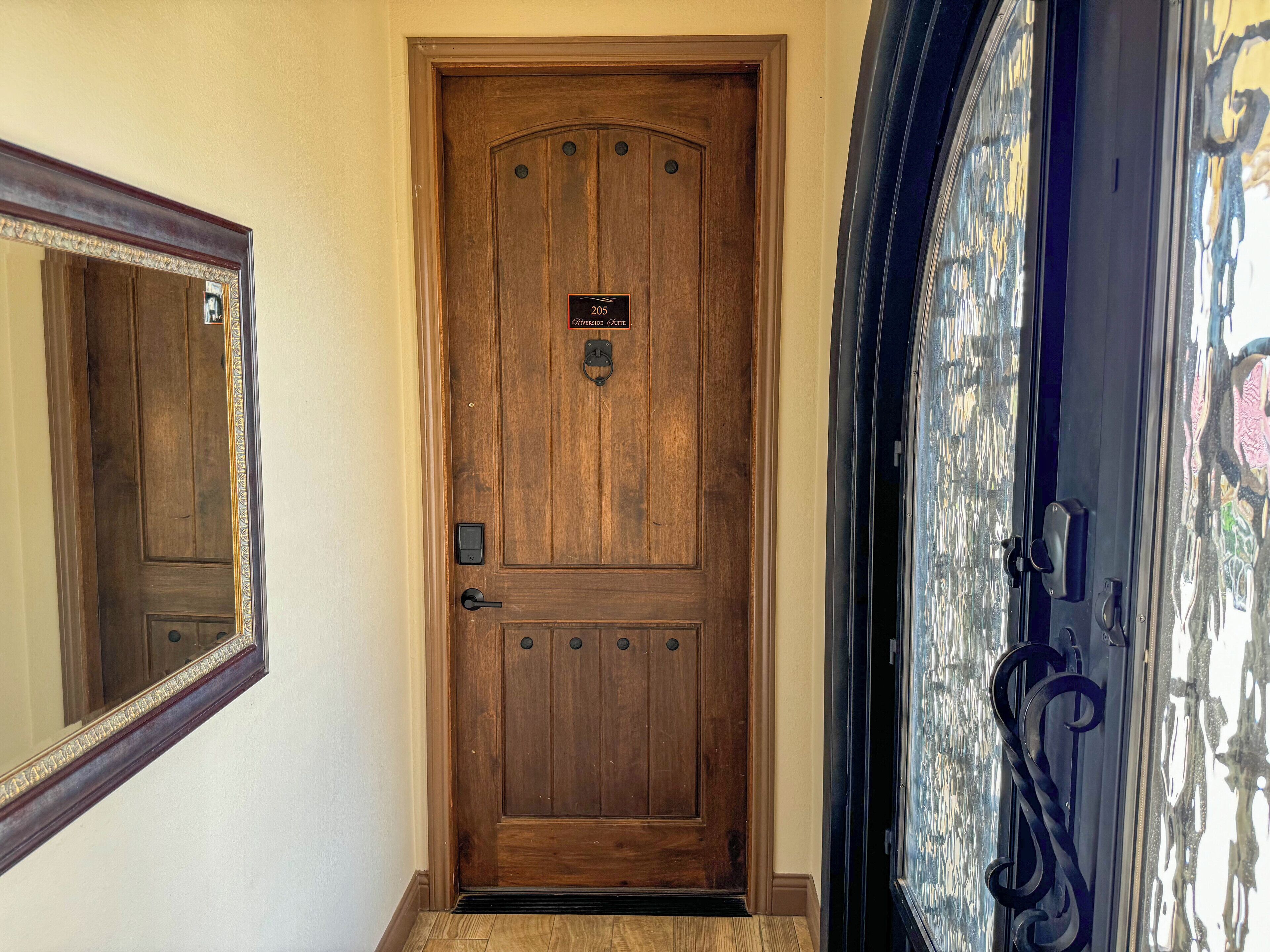 A wooden door with a brass knocker and a plaque, a mirror above it, and a glass door with a metal handle to the right.