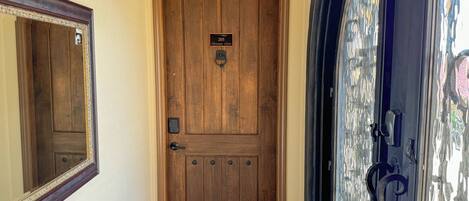 A wooden door with a brass knocker and a plaque, a mirror above it, and a glass door with a metal handle to the right.
