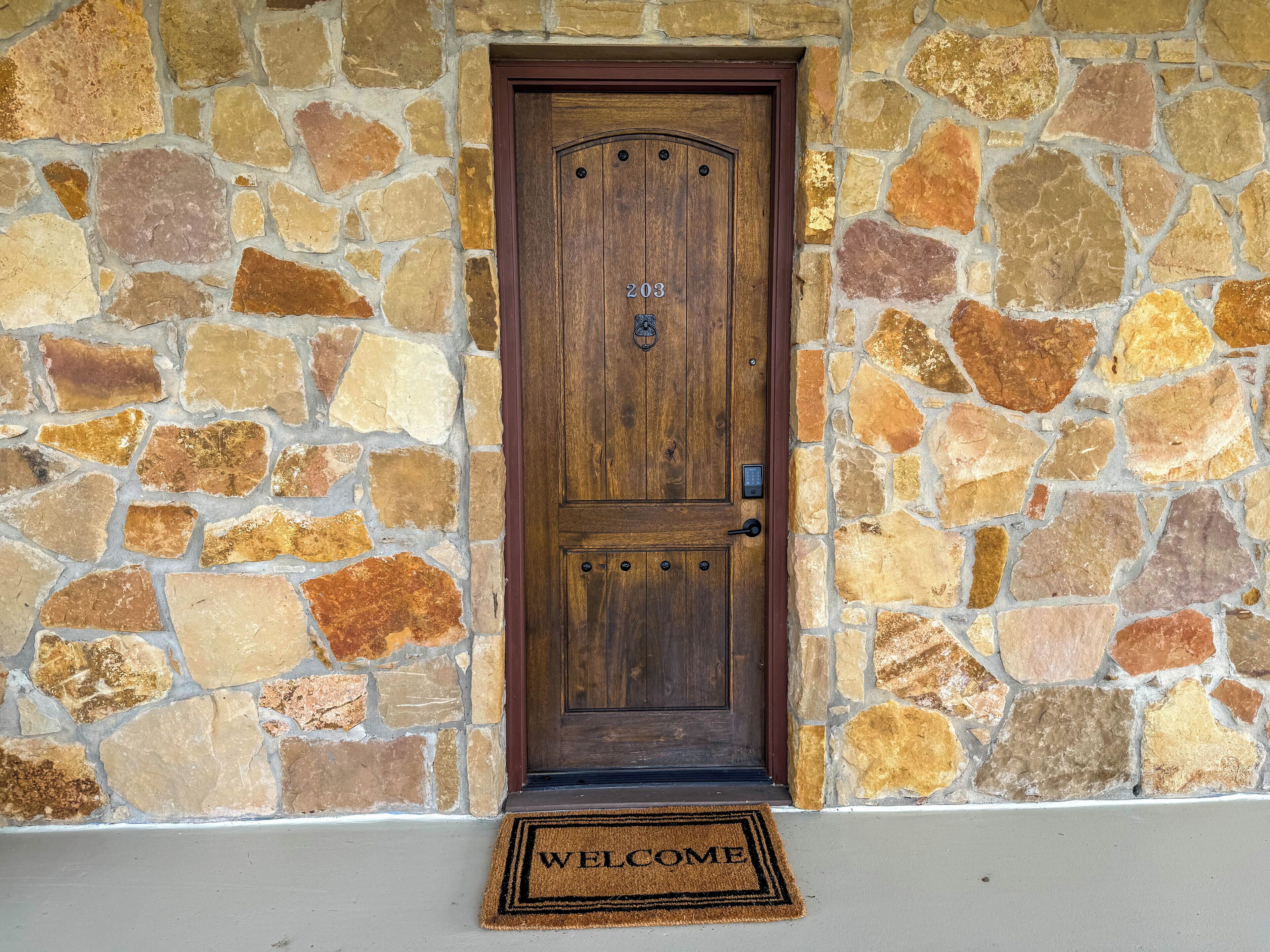 A stone wall with a wooden door marked "203" and a welcome mat.