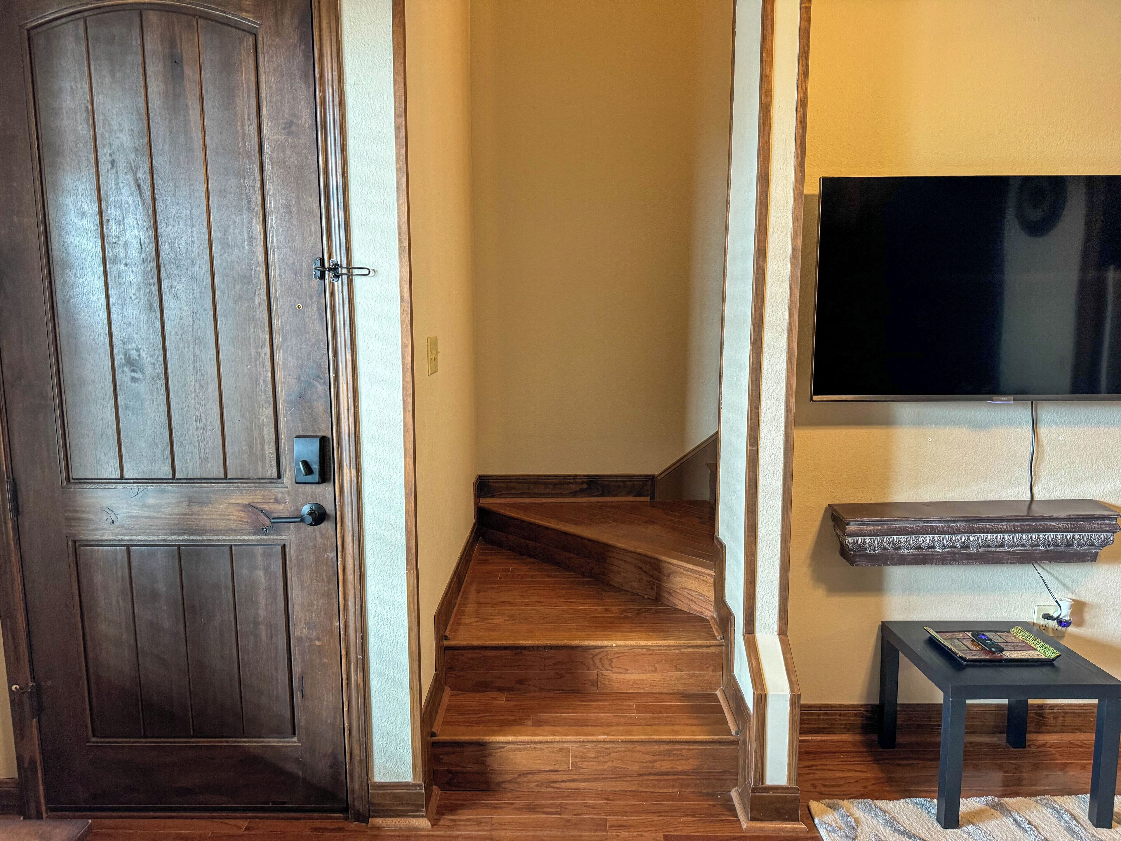 A wooden door, a staircase, a flat-screen TV mounted on the wall, and a small table with a keyboard and mouse.