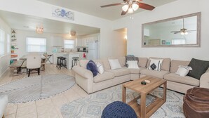 A spacious living area with a large beige sectional sofa, a wooden coffee table, and a ceiling fan. A kitchen is visible in the background with white cabinets and a dining area. - Mai Tide--4 BR House, Sleeps 11, Close to Beach, Dogs OK, Tons of Parking! (Port Aransas)