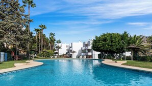 A swimming pool with a clear blue water surface, surrounded by palm trees and a white building with balconies in the background.