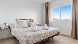 A modern bedroom with a large bed, a bedside table with a lamp, and a view of the sky through a window.