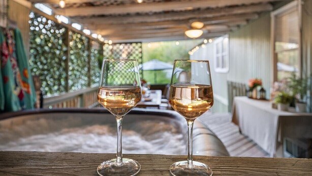Two glasses of white wine on a wooden table in a cozy outdoor seating area with a hot tub in the background. - Hot Tub, Fire pit, Cabana, Covered Patio & Pet-friendly Riverside Retreat (New Braunfels)