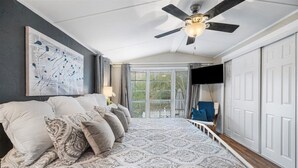 A bedroom with a large bed, a ceiling fan, a TV, and a view of a white fence through the window. - Hot Tub, Fire pit, Cabana, Covered Patio & Pet-friendly Riverside Retreat (New Braunfels)