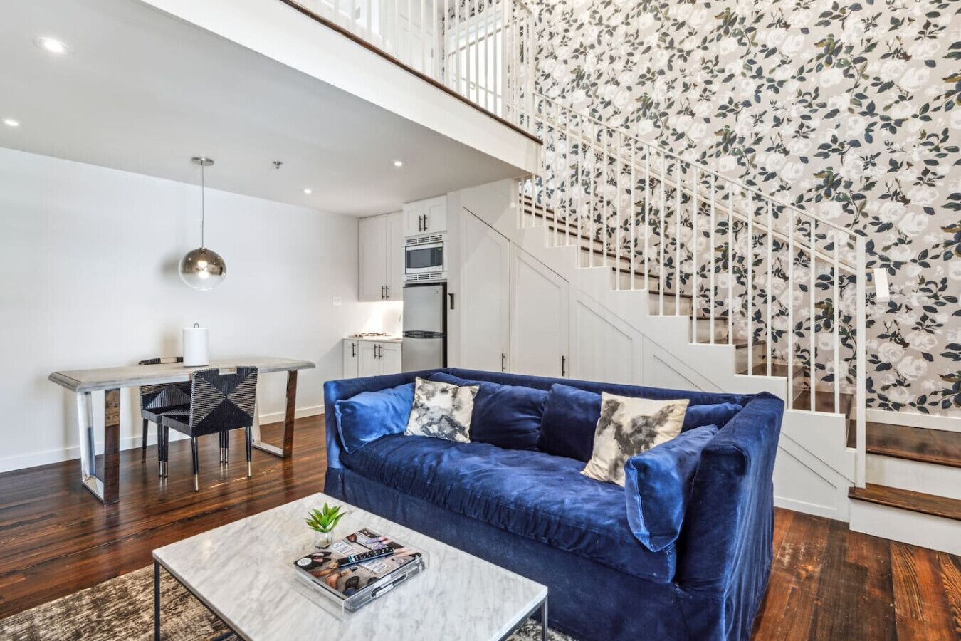 Un salon moderne avec un canapé bleu, une table basse avec des magazines, un coin repas avec une table blanche et un escalier orné d’un papier peint à motifs floraux.