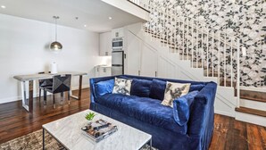A modern living room with a blue sofa, a coffee table with magazines, a dining area with a white table, and a staircase with floral wallpaper. - The Belle on Magazine (New Orleans)