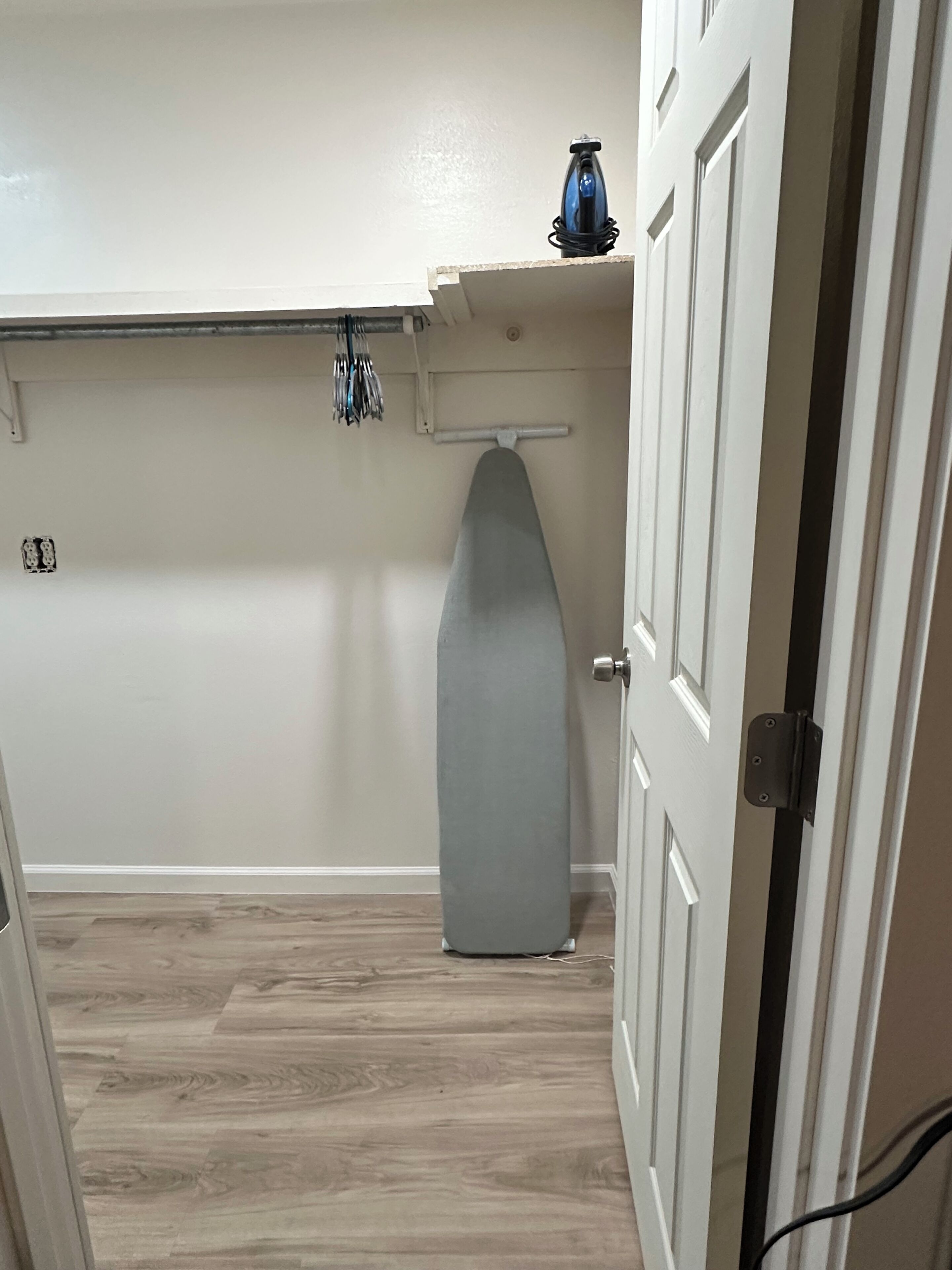 A laundry room with a hanging rack, a large gray ironing board, and a small blue container on a shelf.