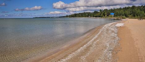 A beach with clear water, a sandy shore, and a house in the background.