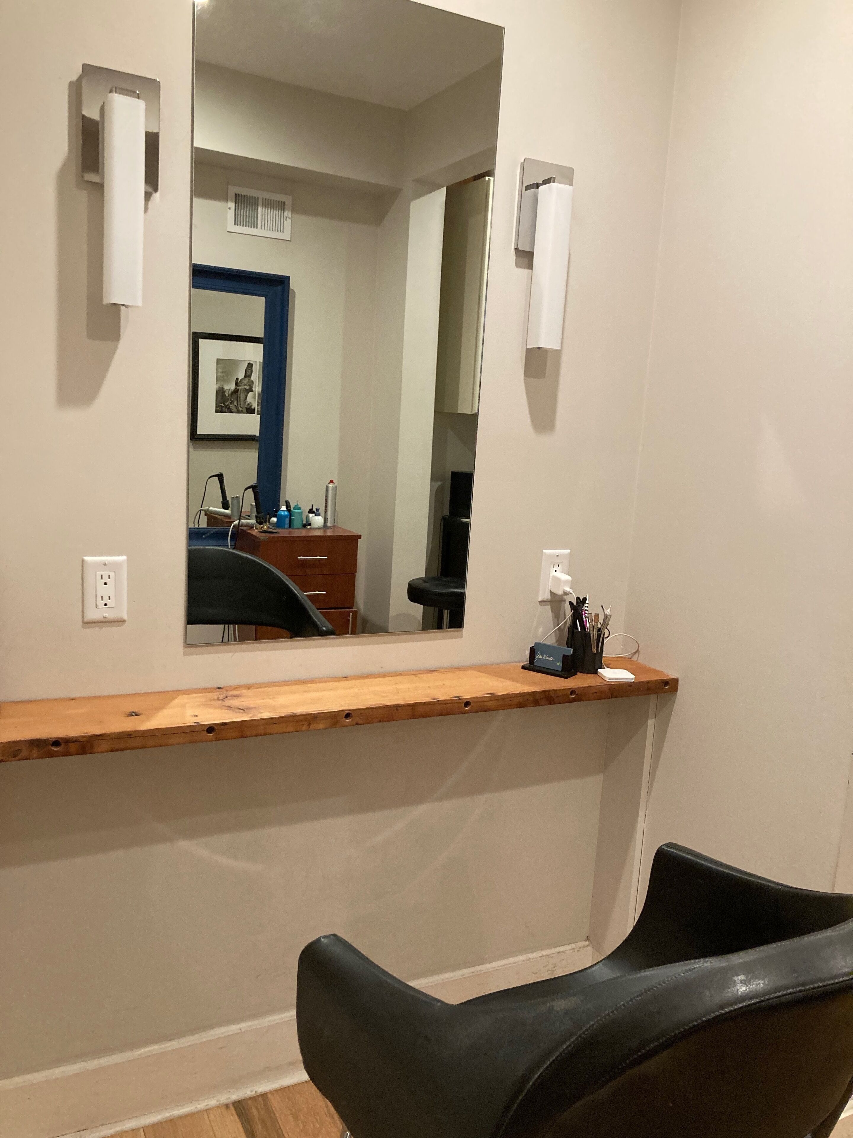 Una habitación con mostrador de madera, una silla negra, un espejo grande y un escritorio pequeño con una computadora y varios objetos.