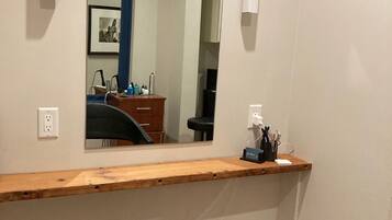 Una habitación con mostrador de madera, una silla negra, un espejo grande y un escritorio pequeño con una computadora y varios objetos.