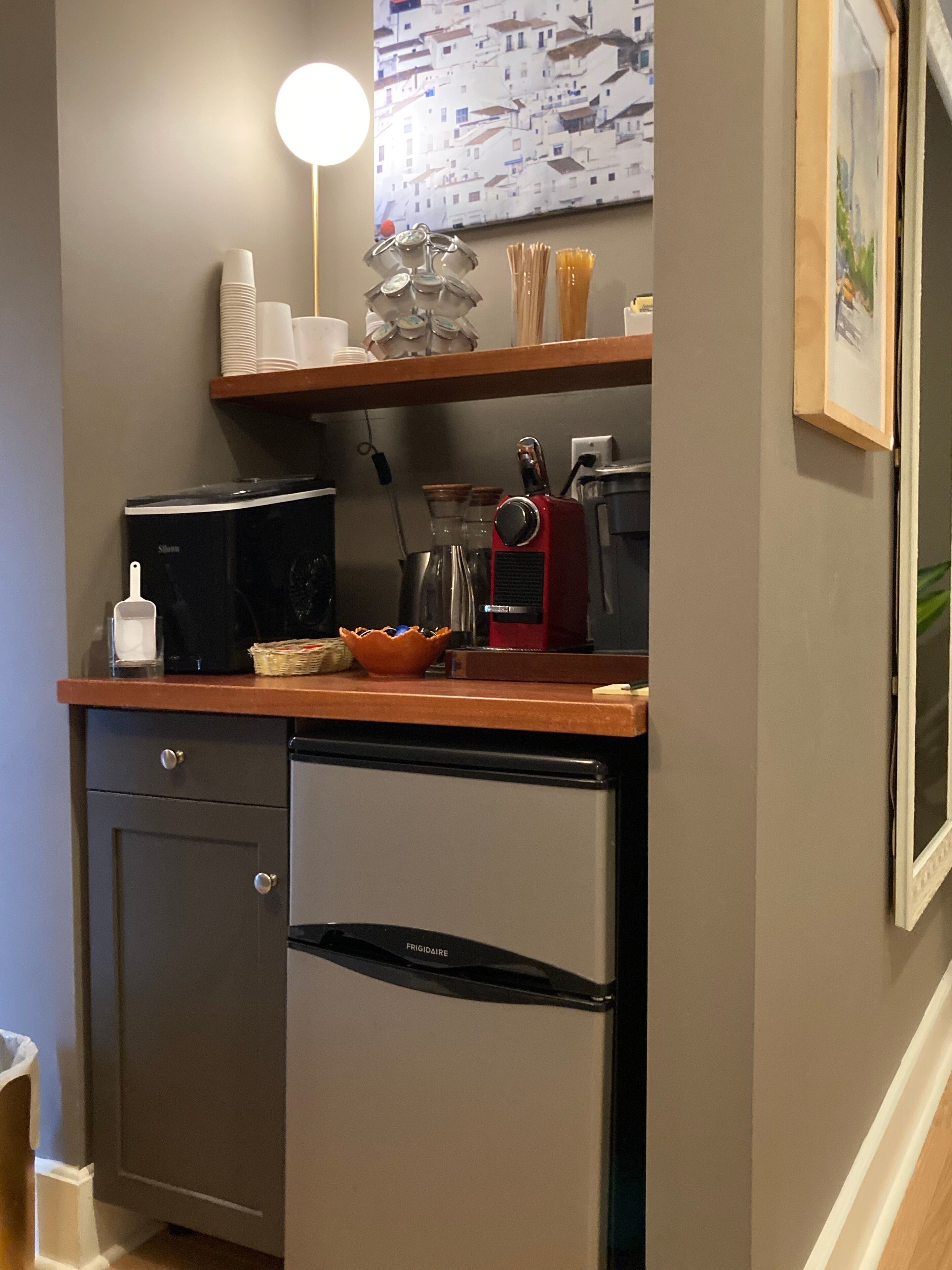 A compact kitchen with a refrigerator, microwave, and coffee maker. There are shelves above the counter holding cups and a painting.