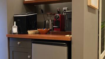A compact kitchen with a refrigerator, microwave, and coffee maker. There are shelves above the counter holding cups and a painting.