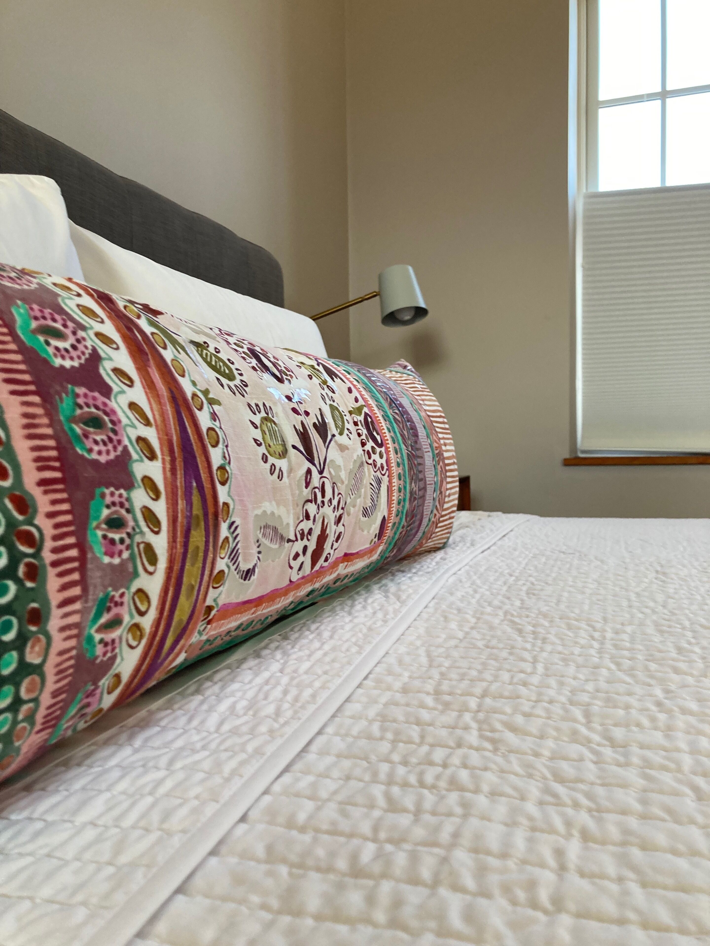 Una cama con una manta y almohadas estampadas, una lámpara de noche y una ventana con persianas.