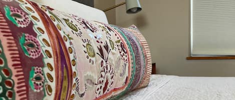 Una cama con una manta y almohadas estampadas, una lámpara de noche y una ventana con persianas.